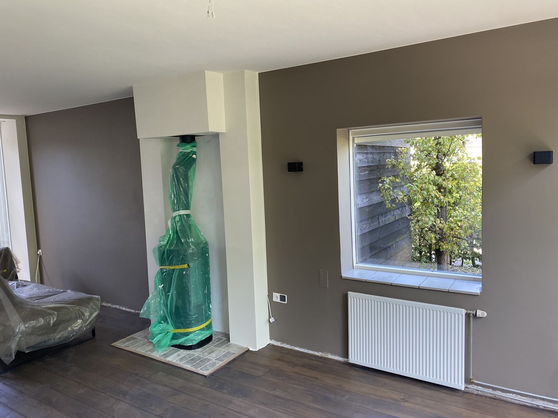 Woonkamer tijdens het schilderwerk: taupe wanden, witte schoorsteen, kachel en bank ingepakt in plastic