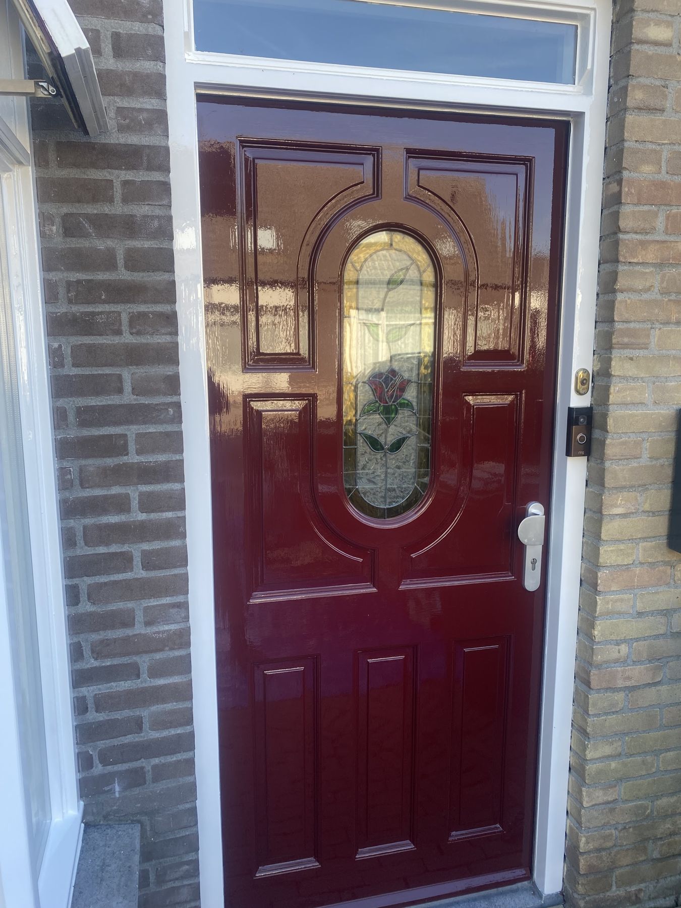 Bordeauxrode voordeur in hoogglans met een ovaal glas-in-loodraam met een roos, in een wit kozijn